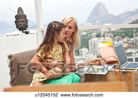 Stock Image - "Mother and daughter sitting at table on balcony, looking through book". Fotosearch