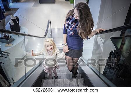 Stock Image - Mother and young daughter moving up escalator in shopping mall. Fotosearch