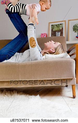 Stock Image - "Mother lying on sofa, holding baby girl in air". Fotosearch
