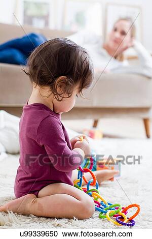 Stock Image - Mother lying on sofa watching baby boy play on floor. Fotosearch