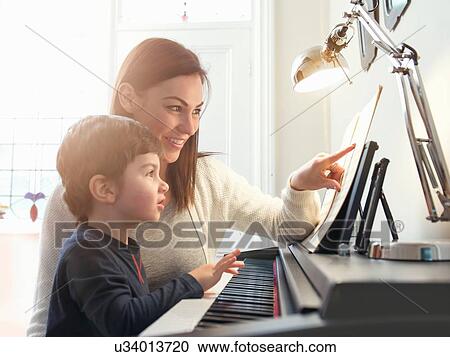 Stock Image - Mother pointing at sheet music to teach son to play piano at home. Fotosearch