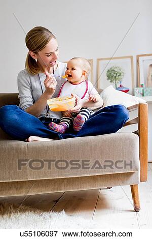 Stock Photo - "Mother sitting on sofa, feeding baby girl". Fotosearch