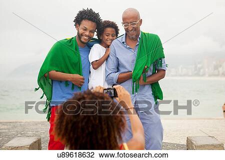 Stock Image - "Over shoulder view of mother photographing family wrapped in Brazilian flag on Ipanema beach, Rio De Janeiro, Brazil". Fotosearch