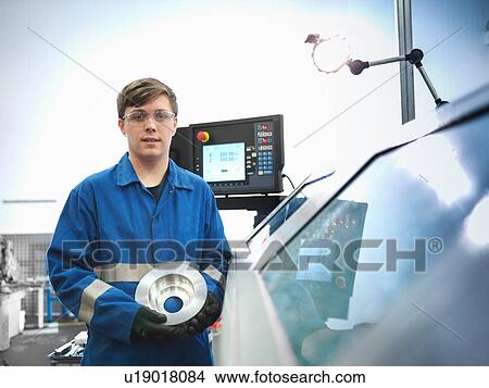Picture - Portrait of apprentice engineer holding steel component with lathe in engineering factory. Fotosearch