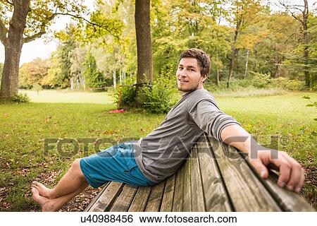Stock Photograph - Portrait of barefoot young man sitting on park bench. Fotosearch