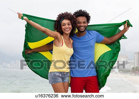 Stock Photograph - "Portrait of couple holding up Brazilian flag on Ipanema beach, Rio De Janeiro, Brazil". Fotosearch