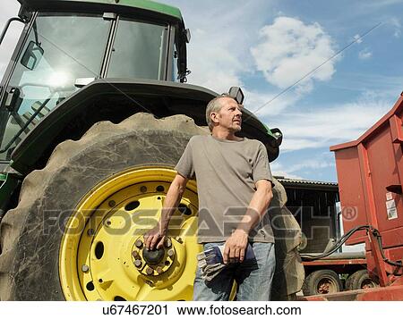 Stock Image - "Portrait of farmer standing next to tractor, looking away". Fotosearch