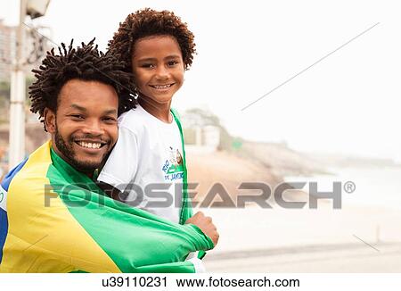 Stock Image - "Portrait of father and son wrapped in Brazilian flag on Ipanema beach, Rio De Janeiro, Brazil". Fotosearch