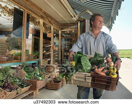 Stock Image - Portrait of male organic farmer with basket of organic vegetables outside farm shop. Fotosearch