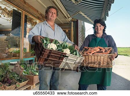 Stock Image - Portrait of organic farming couple with baskets of organic vegetables outside farm shop. Fotosearch