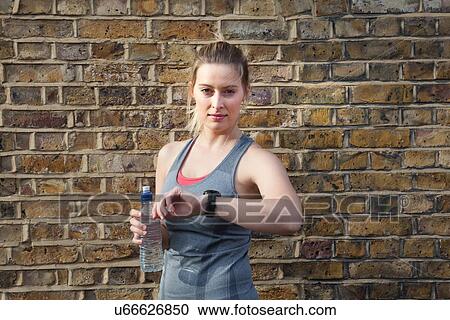 Stock Image - Portrait of young female runner in front of brick wall checking stop watch. Fotosearch