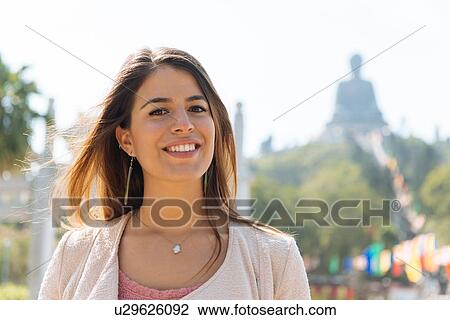 Stock Image - "Portrait of young female tourist in front of Tian Tan Buddha, Po Lin Monastery, Lantau Island, Hong Kong, China". Fotosearch