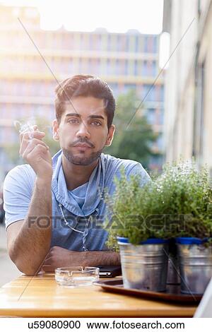 "Portrait of young man outdoors, sitting at table, smoking cigarette" View Large Photo Image Stock Image - "Portrait of young man outdoors, sitting at table, smoking cigarette". Fotosearch