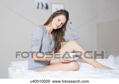 Stock Photo - Portrait of young woman sitting on bed drinking coffee. Fotosearch