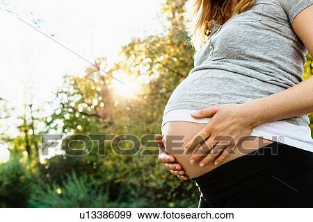 Stock Photo - "Pregnant woman holding belly, outdoors, mid section". Fotosearch