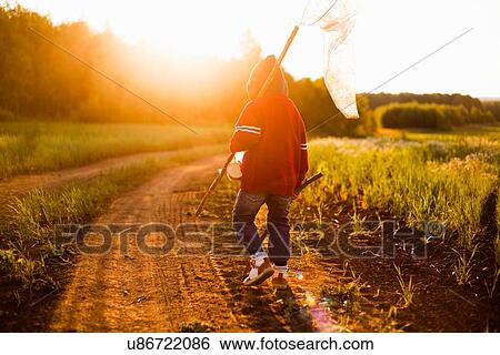 Stock Photograph - "Rear view of boy with butterfly net walking along dirt track at sunset, Sarsy village, Sverdlovsk Region, Russia". Fotosearch