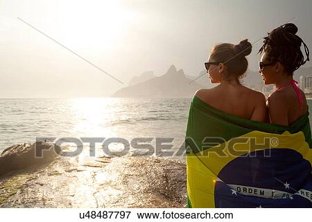 Stock Photo - "Rear view of two young women wrapped in Brazilian flag, Ipanema beach, Rio De Janeiro, Brazil". Fotosearch