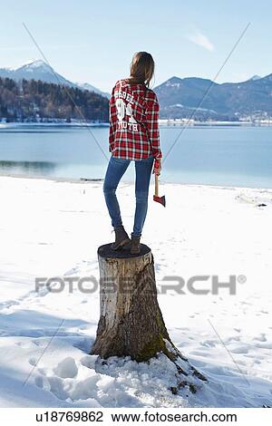 Stock Image - "Rear view of young woman standing on tree stump at snow covered lakeside, German Alps, Germany". Fotosearch