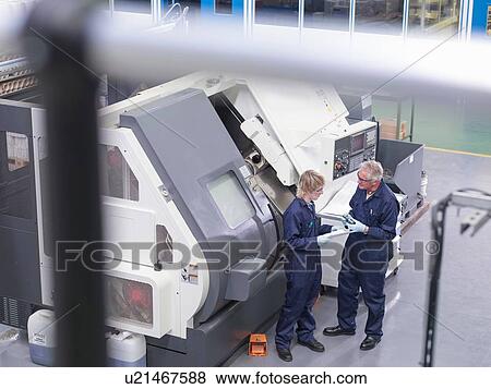 Stock Photo - "Senior engineer instructing apprentice on factory machinery, high angle view". Fotosearch