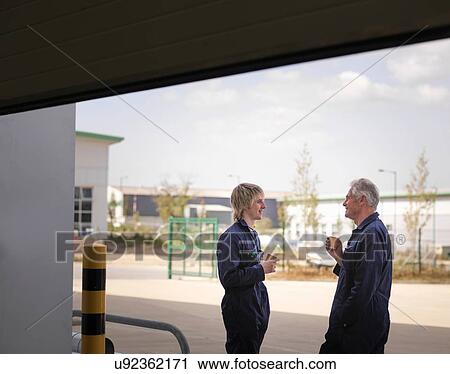 Stock Image - Senior engineer taking a coffee break with apprentice in factory doorway. Fotosearch