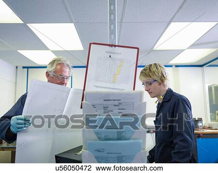 Stock Image - Senior engineer with blueprint instructing apprentice in factory. Fotosearch