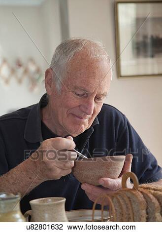 Stock Image - "Senior man, sitting at table, eating bowl of cereal". Fotosearch