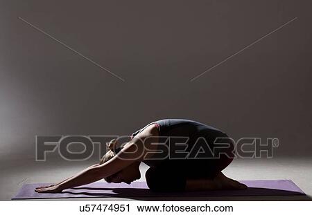 Stock Image - Silhouetted low key shot of young woman practicing yoga bending forward on mat. Fotosearch
