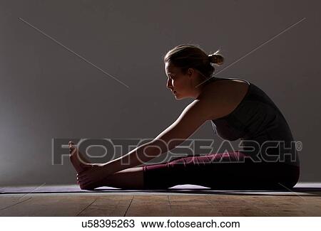 Stock Image - Silhouetted low key shot of young woman practicing yoga touching toes on mat. Fotosearch