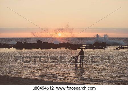 Stock Image - "Silhouetted mother and female toddler paddling in sea looking out at waves splashing, Kona, Hawaii, USA". Fotosearch