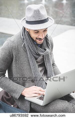 Stock Image - Smiling businessman sitting on wall using laptop. Fotosearch