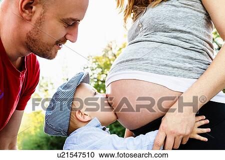 Stock Image - "Son kissing mum's pregnant belly while father looks on, mid section". Fotosearch