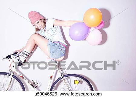 Stock Photograph - Studio shot of young woman with feet up on bicycle holding bunch of balloons. Fotosearch