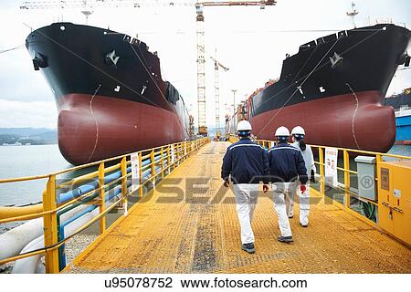 Stock Image - "Three workers walking towards ships at shipping port, rear view, GoSeong-gun, South Korea". Fotosearch