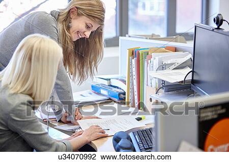 Stock Image - Two businesswomen checking paperwork at office desk. Fotosearch