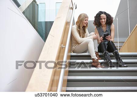 Two businesswomen sitting on office stairway reading smartphone texts View Large Photo Image Stock Image - Two businesswomen sitting on office stairway reading smartphone texts. Fotosearch