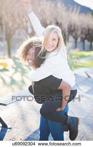 Two female friends giving piggy backs in park View Large Photo Image Picture - Two female friends giving piggy backs in park. Fotosearch
