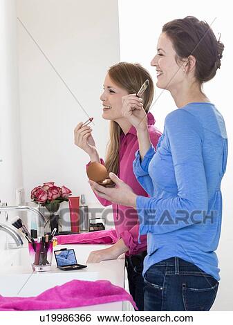 Stock Photograph - Two female friends gossiping and applying lipstick in bathroom. Fotosearch