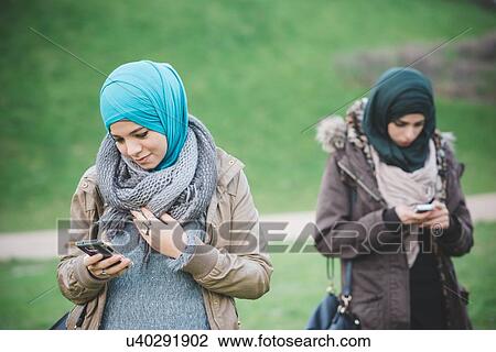 Stock Image - Two female friends in park reading texts on smartphones. Fotosearch