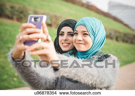 Picture - Two female friends in park taking smartphone selfie. Fotosearch