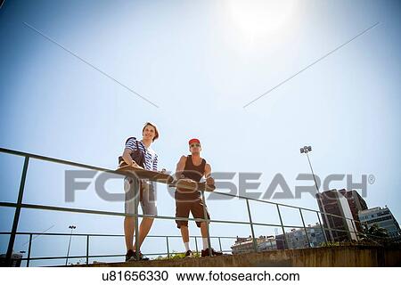 "Two men, holding skateboards, leaning on railings, low angle view" View Large Photo Image Stock Image - "Two men, holding skateboards, leaning on railings, low angle view". Fotosearch