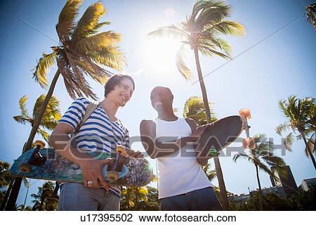 Stock Image - "Two men, holding skateboards, walking outdoors". Fotosearch