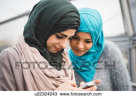 Stock Image - Two young female friends reading smartphone texts on park path. Fotosearch