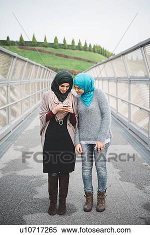Stock Photography - Two young female friends reading smartphone texts on park footbridge. Fotosearch