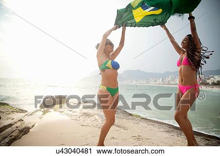 Stock Image - "Two young women dancing whilst holding up Brazilian flag, Ipanema beach, Rio De Janeiro, Brazil". Fotosearch