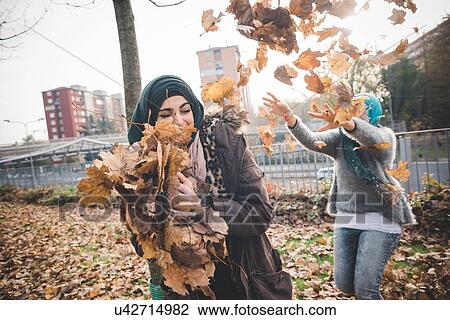 Stock Image - Two young women friends play fighting with autumn leaves in park. Fotosearch