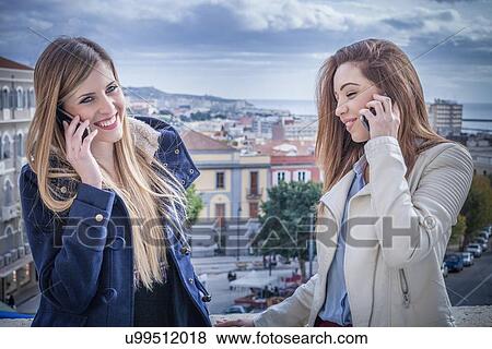 Stock Photo - "Two young women on rooftop talking on smartphones, Cagliari, Sardinia, Italy". Fotosearch