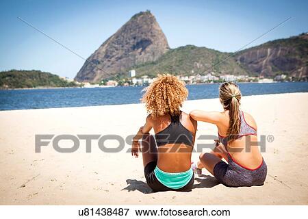 Stock Photo - "Two young women sitting on beach, Rio de Janeiro, Brazil". Fotosearch