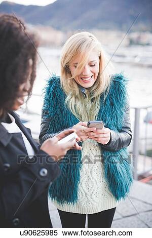 Stock Photography - "Two young women texting on smartphones at Lake Como, Como, Italy". Fotosearch