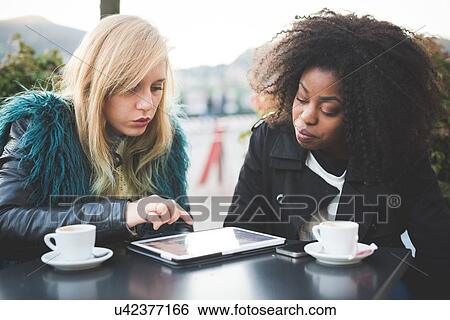 Stock Photograph - "Two young women using touchscreen on digital tablet at sidewalk cafe, Lake Como, Como, Italy". Fotosearch