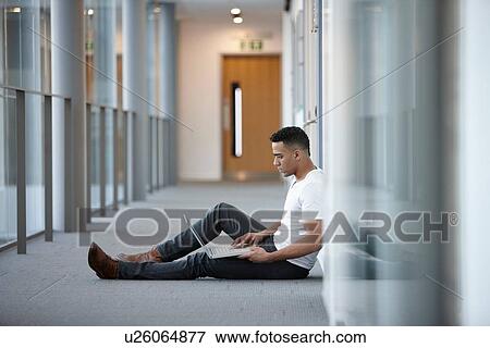 Stock Photo - Young businessman sitting in office corridor using laptop. Fotosearch